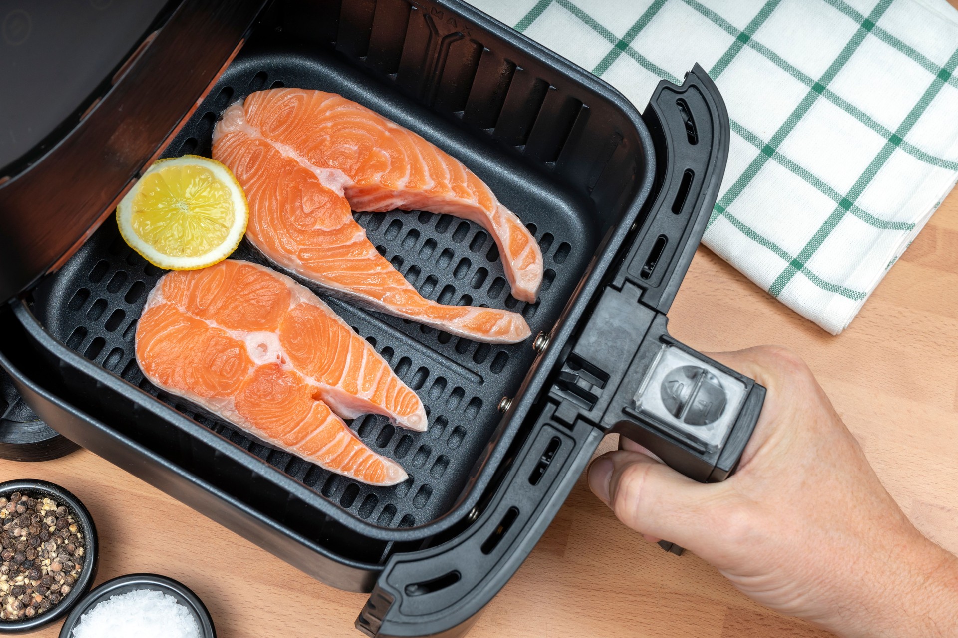 HAND HOLDING AIR FRYER BASKET WITH TWO SLICES OF RAW SALMON READY TO COOK IN THE KITCHEN. TOP VIEW.